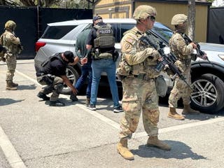 Three heavily armed National Guard troops are visible two people are detained next to a vehicle in a parking lot